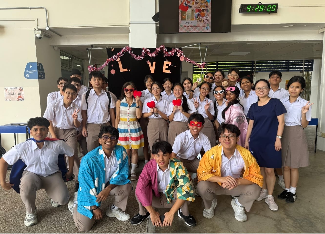 A class of students and two teachers pose cheerfully in front of a decorated "L.V.E" backdrop, some wearing fun props like heart-shaped sunglasses and colourful jackets.